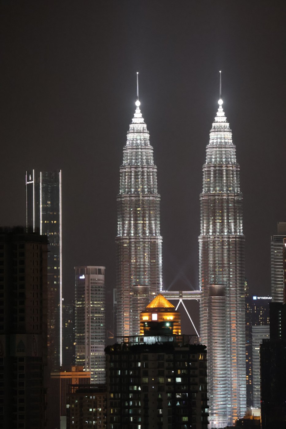 The Petronas Towers lit white against a hazy night sky, with the Kuala Lumpur skyline below