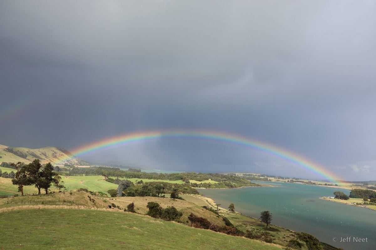 A rainbow arcs over a grey-blue inlet and green hills, with dark storm clouds above.