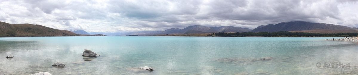 Several rocks break the surface of still grey-blue water, with low hills and cloud reflected in the lake.