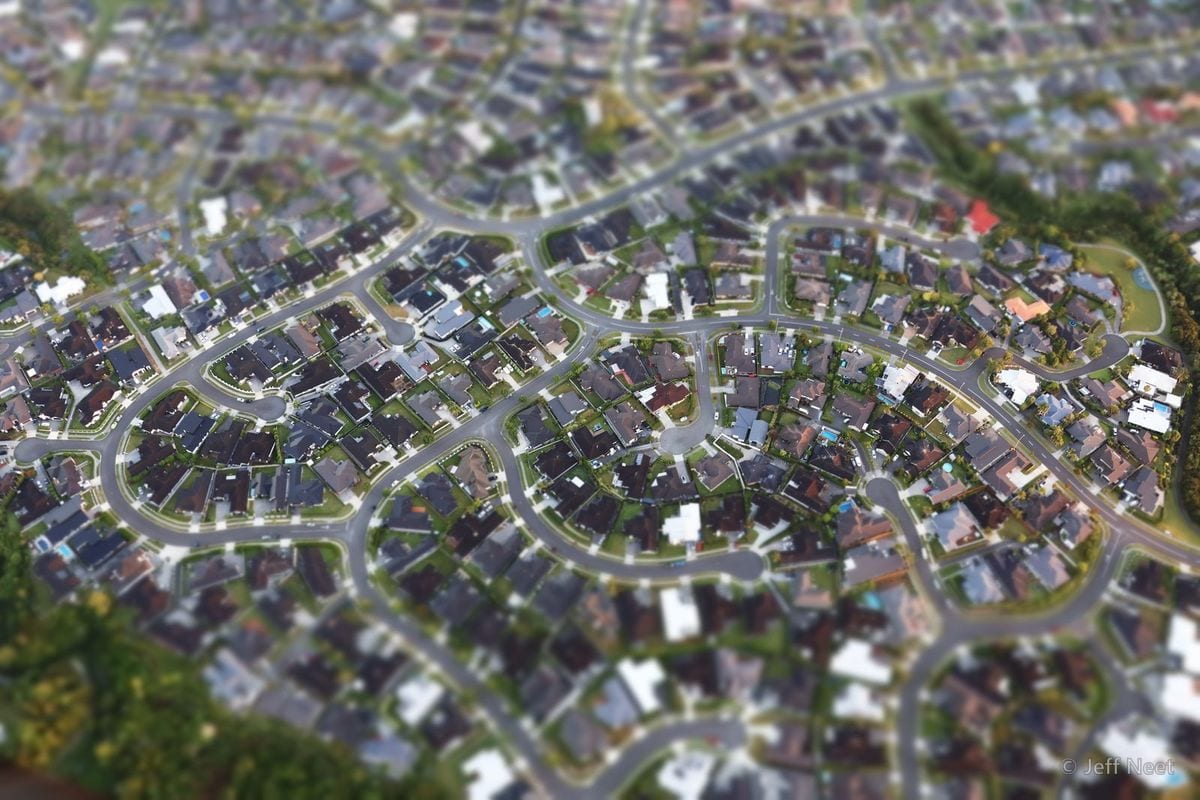 An aerial view of a suburban housing estate with curving streets, rooftops and trees, blurred at the edges.
