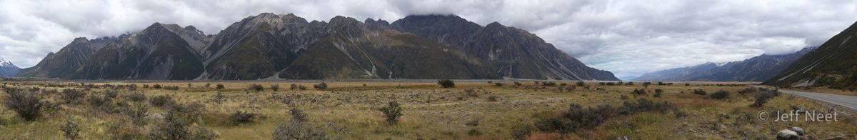 Wide panoramic view of grey mountain ridges under low cloud, with dry tussock grass across the valley floor.