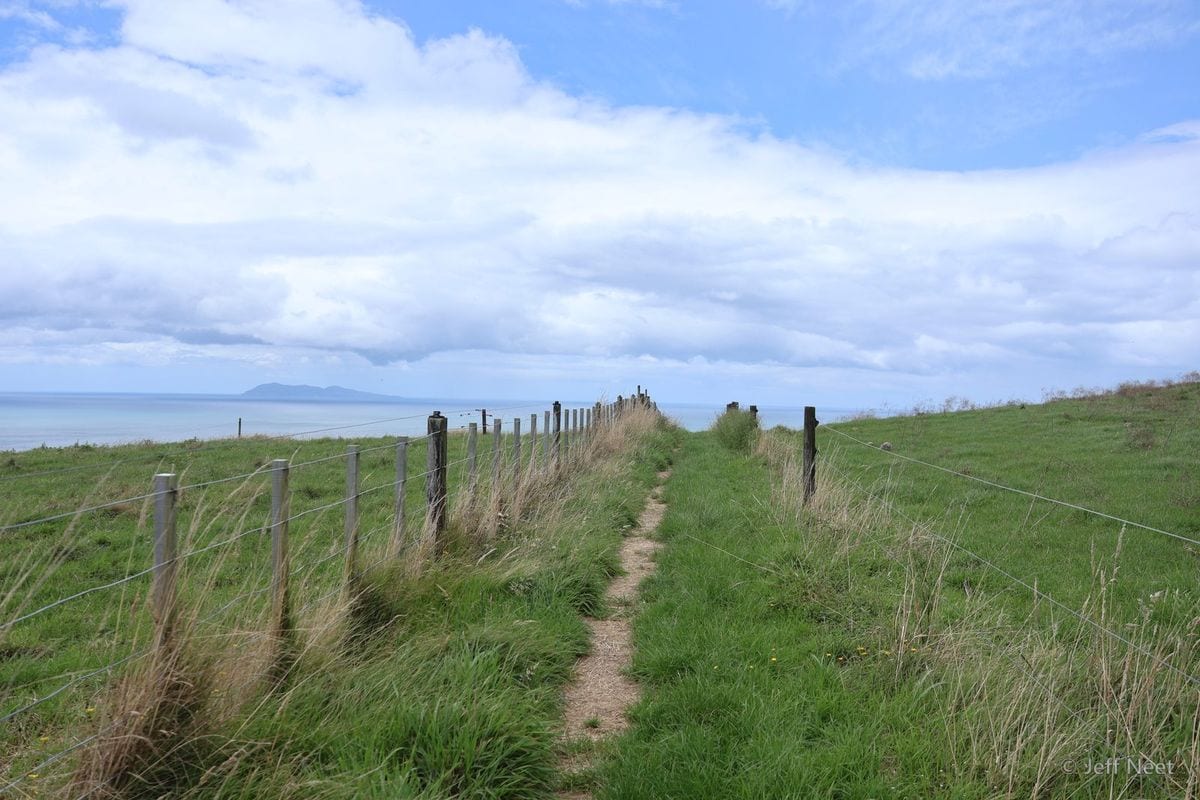 A single distant figure walks along a green clifftop beside a fence line above a calm grey-blue sea.