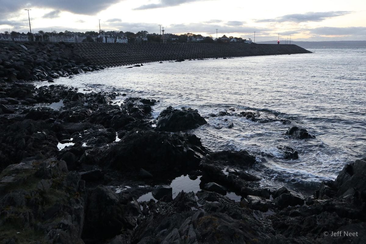 Dark wet rocks and a low sea wall run along the shoreline, with waves washing in under a grey evening sky.