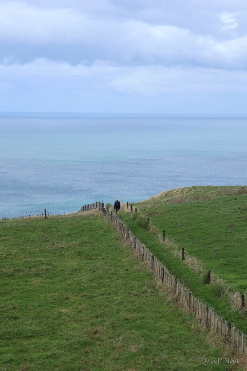 A grassy path runs between wooden fence posts toward the horizon, with pale sea and distant hills under a cloudy sky.