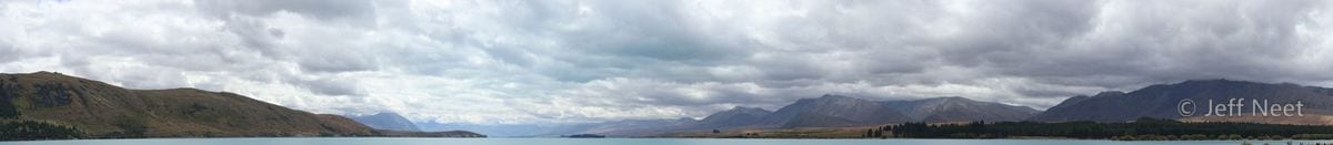 A wide panorama of grey-blue lake and distant mountains under heavy cloud, with dark hills at the left edge.