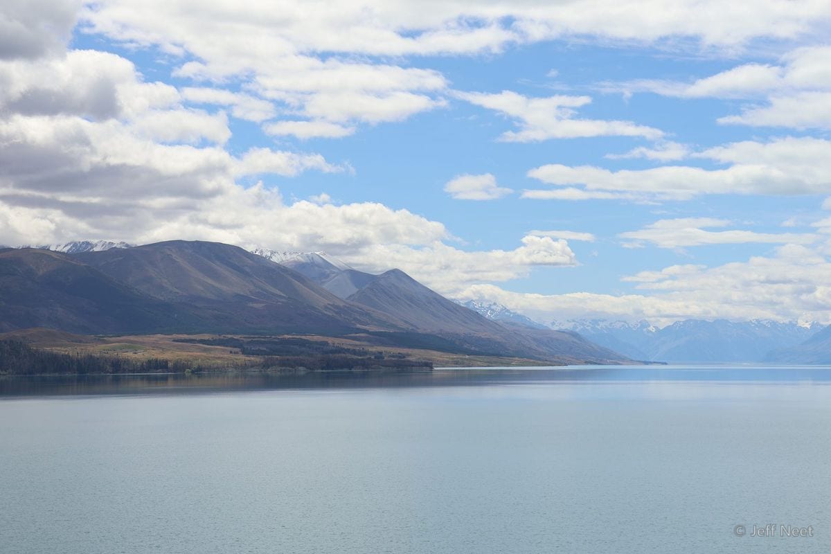 Brown hills and snow-capped ridges recede across a calm grey lake beneath a blue sky with white cloud.