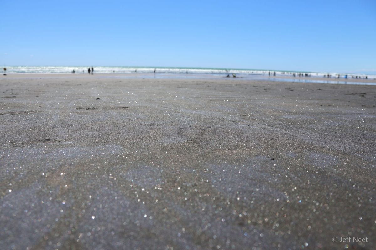 Wet grey-brown sand glitters in the foreground under a clear blue sky, with tiny distant figures at the surf line.