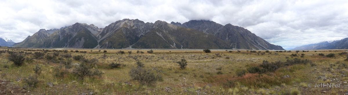 Panoramic view of a long mountain range with cloud over the peaks, above a dry grassy valley floor.