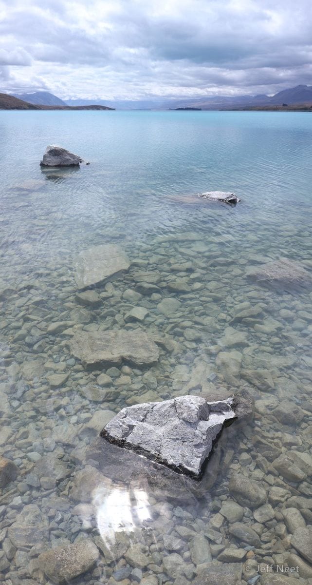 A wide turquoise lake with stones visible through clear shallows, a dark shoreline and distant mountains under cloud.