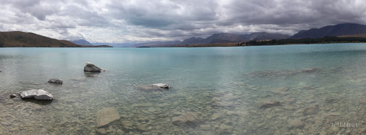 Grey rocks sit in clear turquoise shallows, with a distant shoreline and hills under a cloudy sky.