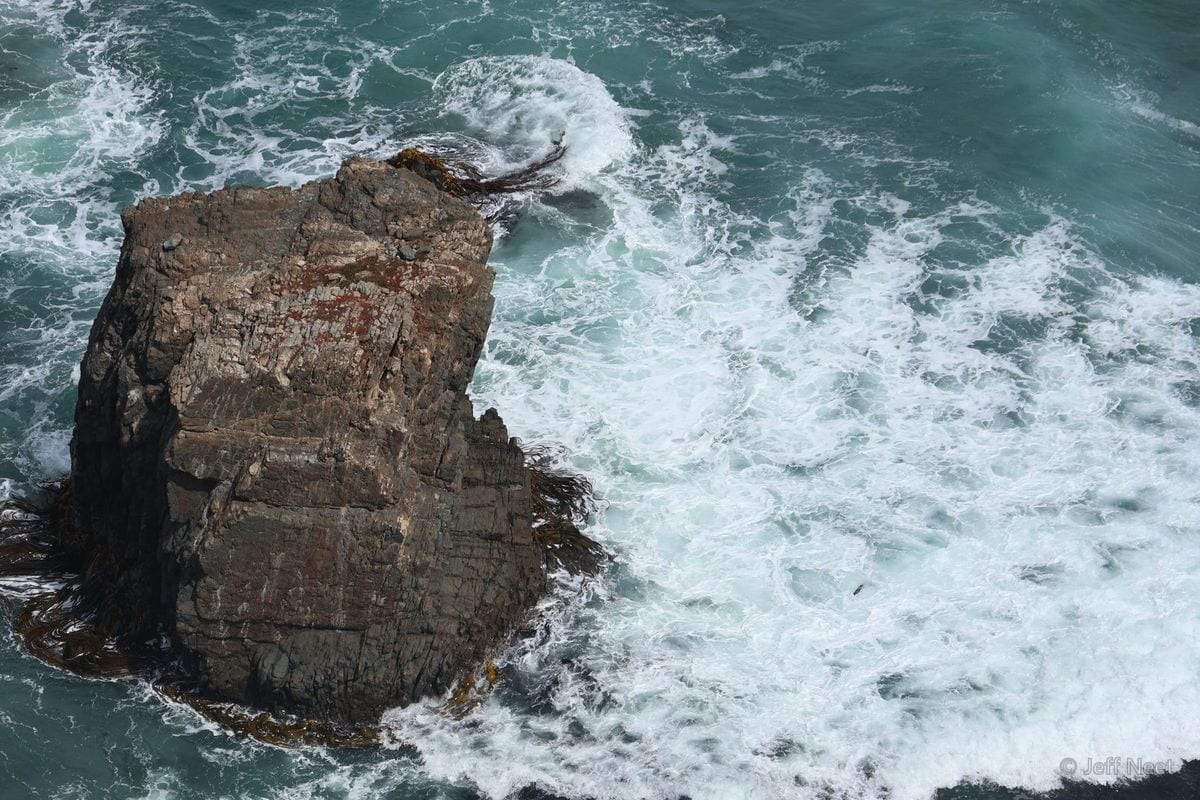 Tall dark rock stack rises from churning white surf in teal water, seen from above.