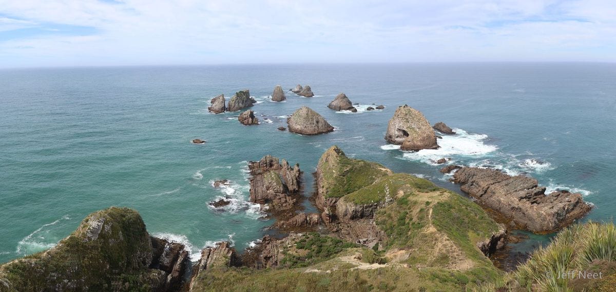 Wide view of offshore rock stacks with white surf around them, above a green rocky headland in soft daylight.