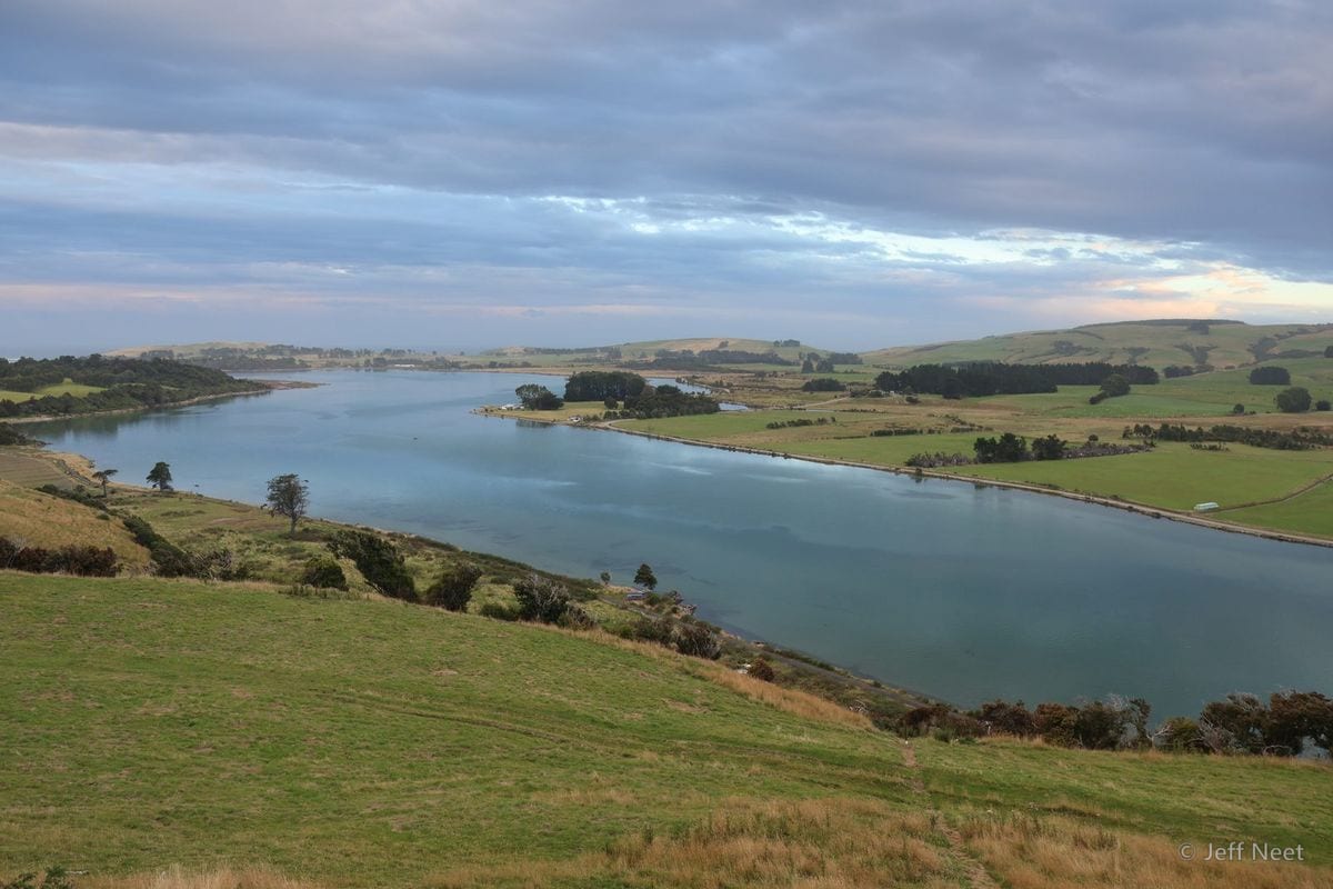 Pink-tinged clouds hang over calm water at dusk, with green hills and dark trees along the shore.