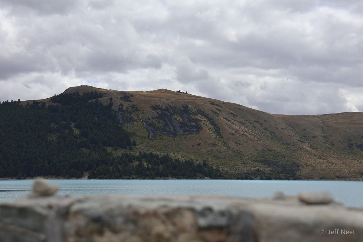 A rough stone wall in the foreground overlooks pale blue water, with a dark conifer-covered hill and tan slopes beyond.