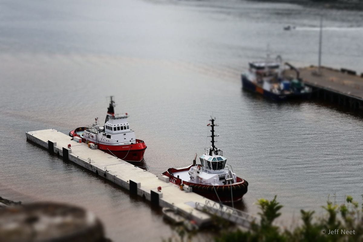 Two red-and-white tugboats are moored beside a grey jetty on calm water, with blurred edges.