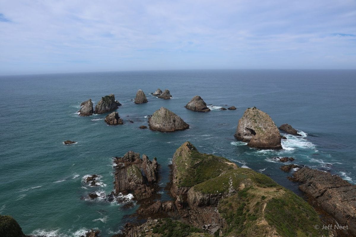 Rock stacks rise from calm teal sea under pale blue sky, with a grassy headland in the foreground.