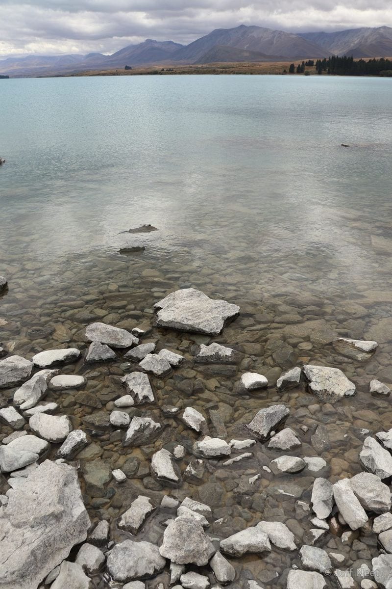 Pale grey rocks lie scattered in clear turquoise shallow water beside a shoreline, with distant hills under cloud.