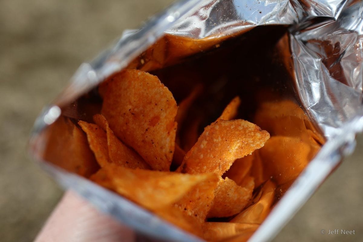 Torn silver foil crisp bag open to show orange crisps, held in a hand against a blurred ground.