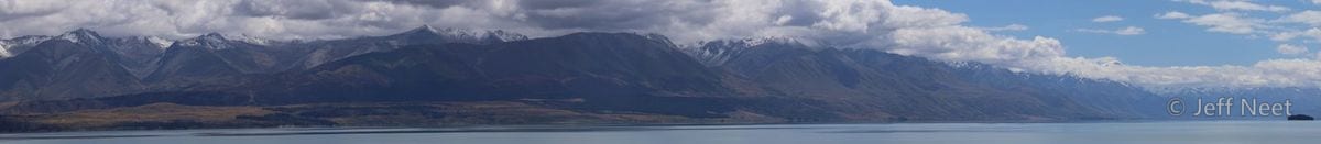 A thin panorama of snow-dusted mountains rises above a grey-blue lake under heavy cloud.