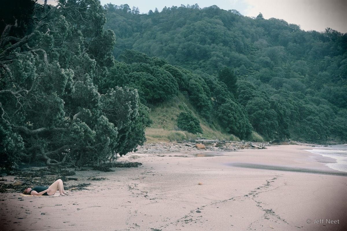 A figure lies on pale sand beside dark-green trees and a steep forested hill by the shore.