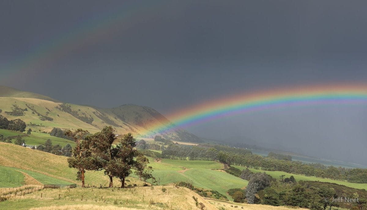 A bright rainbow arcs over blue-green water and green hills under a dark grey sky.