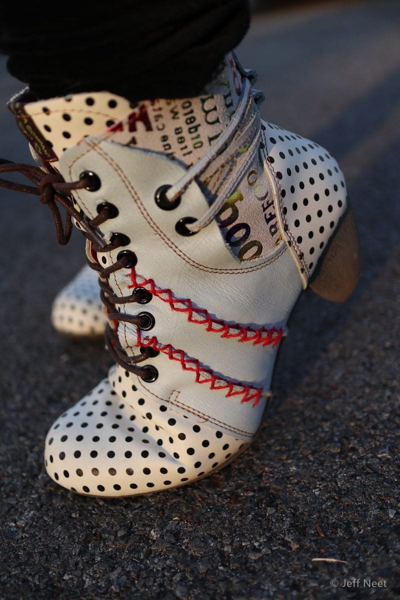Cream leather boot with black polka dots and red stitching stands on dark tarmac, with dark trousers above.