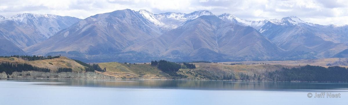Tan and brown hills with dark pine stands rise above a still blue lake, with snowy peaks faint in the distance.