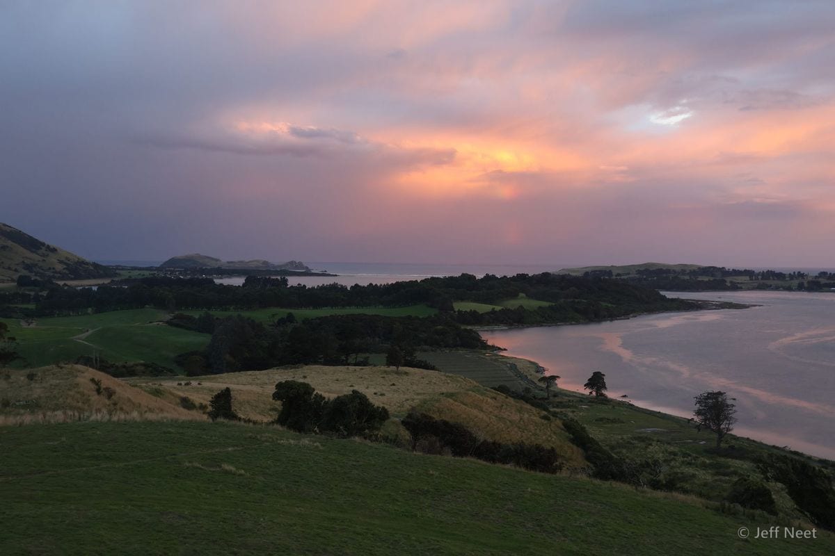 Pink and orange clouds glow above a calm inlet at sunset, with green hills and a dark tree line on the shore.