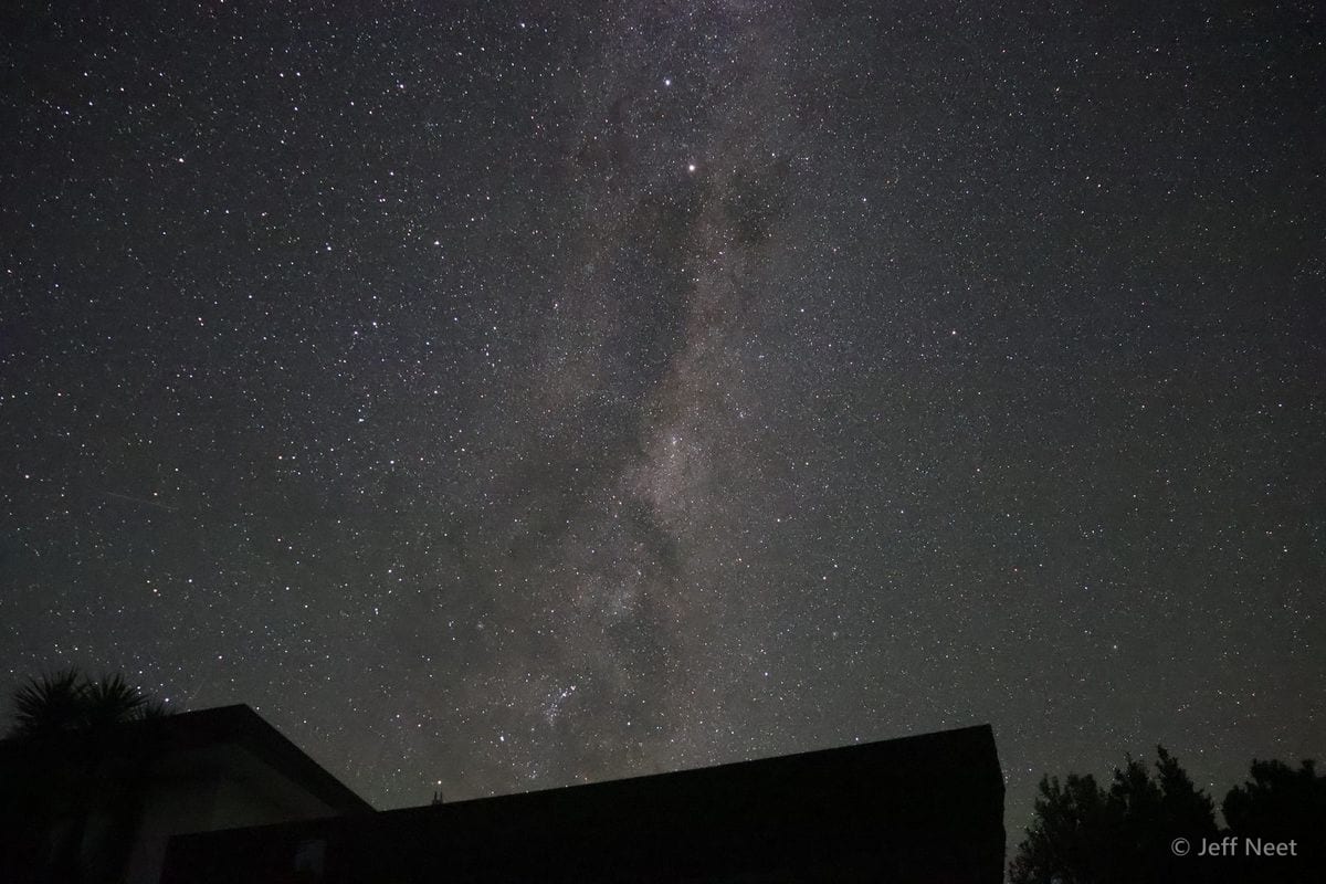 Dense starfield with a faint band of the Milky Way rising above a dark rooftop silhouette at night.