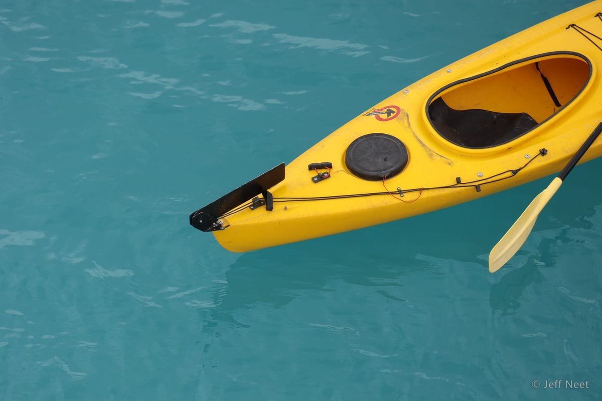 A yellow kayak with a paddle floats on bright turquoise water, its bow pointing to the lower left.